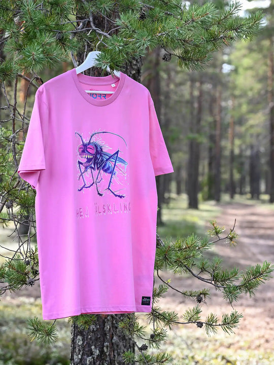 Ein NORRGANIC Unisex T-Shirt aus 100 % Bio-Baumwolle in der Farbe Kaugummipink. Auf der Brust ist ein bunter Mücken-Aufdruck und die Worte HEJ ÄLSKLING (Hey Liebling übersetzt aus dem Schwedischen). Veredelt mit einem Weblabel am unteren Saum und einem kratz-freien Logodruck im Nacken. Normale Passform. T-Shirt hängt an einem Ast. Fotografiert im Wald von Kalix, Schwedisch Lappland.