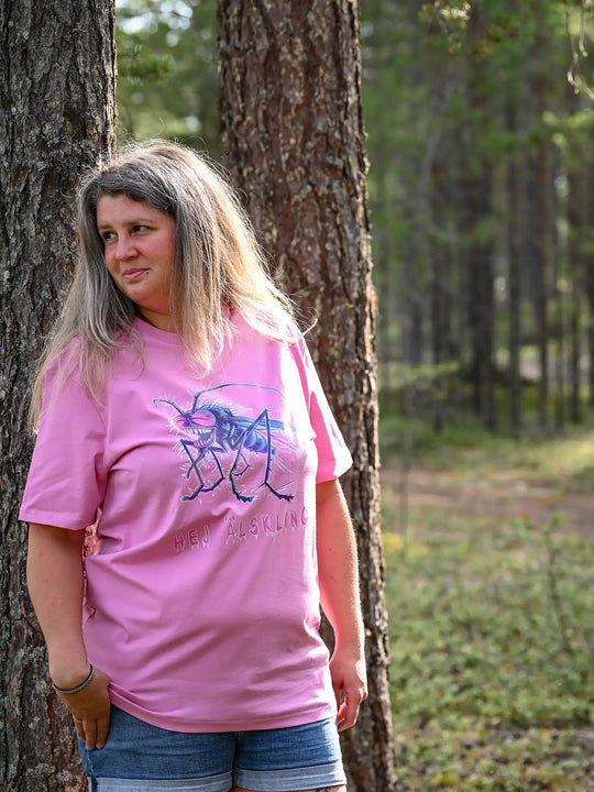 Frau trägt ein NORRGANIC Unisex T-Shirt aus 100 % Bio-Baumwolle in der Farbe Kaugummipink. Auf der Brust ist ein bunter Mücken-Aufdruck und die Worte HEJ ÄLSKLING (Hey Liebling übersetzt aus dem Schwedischen). Veredelt mit einem Weblabel am unteren Saum und einem kratz-freien Logodruck im Nacken. Normale Passform. Fotografiert im Wald von Kalix, Schwedisch Lappland.