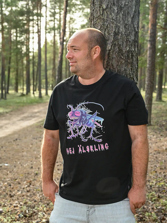 Mann trägt ein schwarzes NORRGANIC Unisex T-Shirt aus 100 % Bio-Baumwolle. Auf der Brust ist ein bunter Mücken-Aufdruck und die Worte HEJ ÄLSKLING (Hey Liebling übersetzt aus dem Schwedischen). Veredelt mit einem Weblabel am unteren Saum und einem kratz-freien Logodruck im Nacken. Normale Passform. Fotografiert im Wald von Kalix, Schwedisch Lappland.