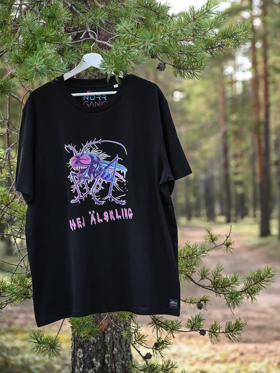 Ein schwarzes NORRGANIC Unisex T-Shirt aus 100 % Bio-Baumwolle. Auf der Brust ist ein bunter Mücken-Aufdruck und die Worte HEJ ÄLSKLING ( Hey Liebling übersetzt aus dem Schwedischen). Veredelt mit einem Weblabel am unteren Saum und einem kratz-freien Logodruck im Nacken. Normale Passform. Das T-Shirt hängt an einem Ast. Fotografiert im Wald von Kalix, Schwedisch Lappland.