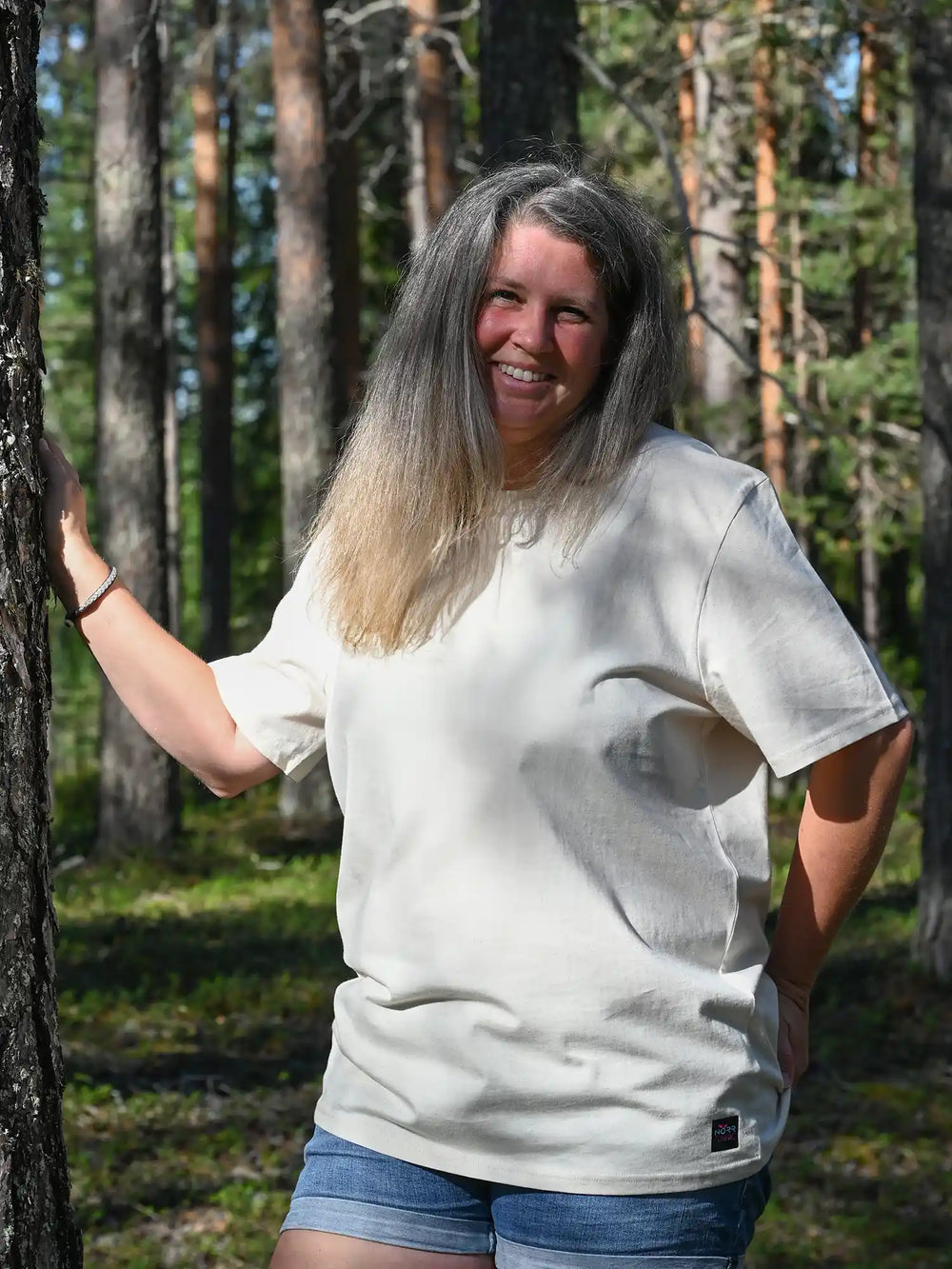 Frau trägt ein naturfarbenes Unisex T-Shirt von NORRGANIC aus 100 % Bio-Baumwolle mit sichtbaren Fasern der Baumwollpflanze. Veredelt mit einem Weblabel am unteren Saum und einem kratz-freien Logodruck im Nacken. Normale Passform. Fotografiert im Wald von Nordschweden.