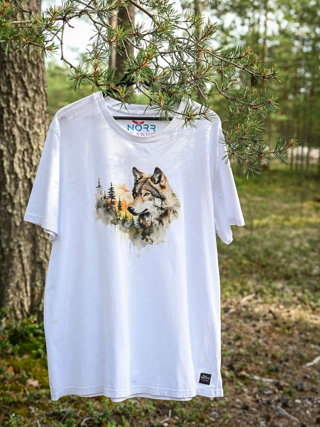 Ein weißes NORRGANIC Unisex T-Shirt aus 100 % Bio-Baumwolle mit einem Wolf-Aufdruck auf der Brust. Veredelt mit einem Weblabel am unteren Saum und einem kratz-freien Logodruck im Nacken. Normale Passform. T-Shirt hängt an einem Ast im Wald von Nordschweden.